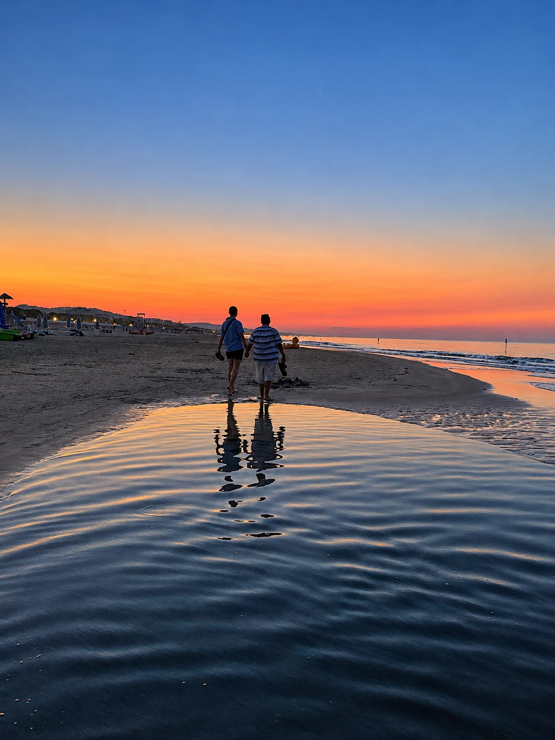 Two people walking along a sandy shoreline beneath a sunset sky, reflected in a pool of water