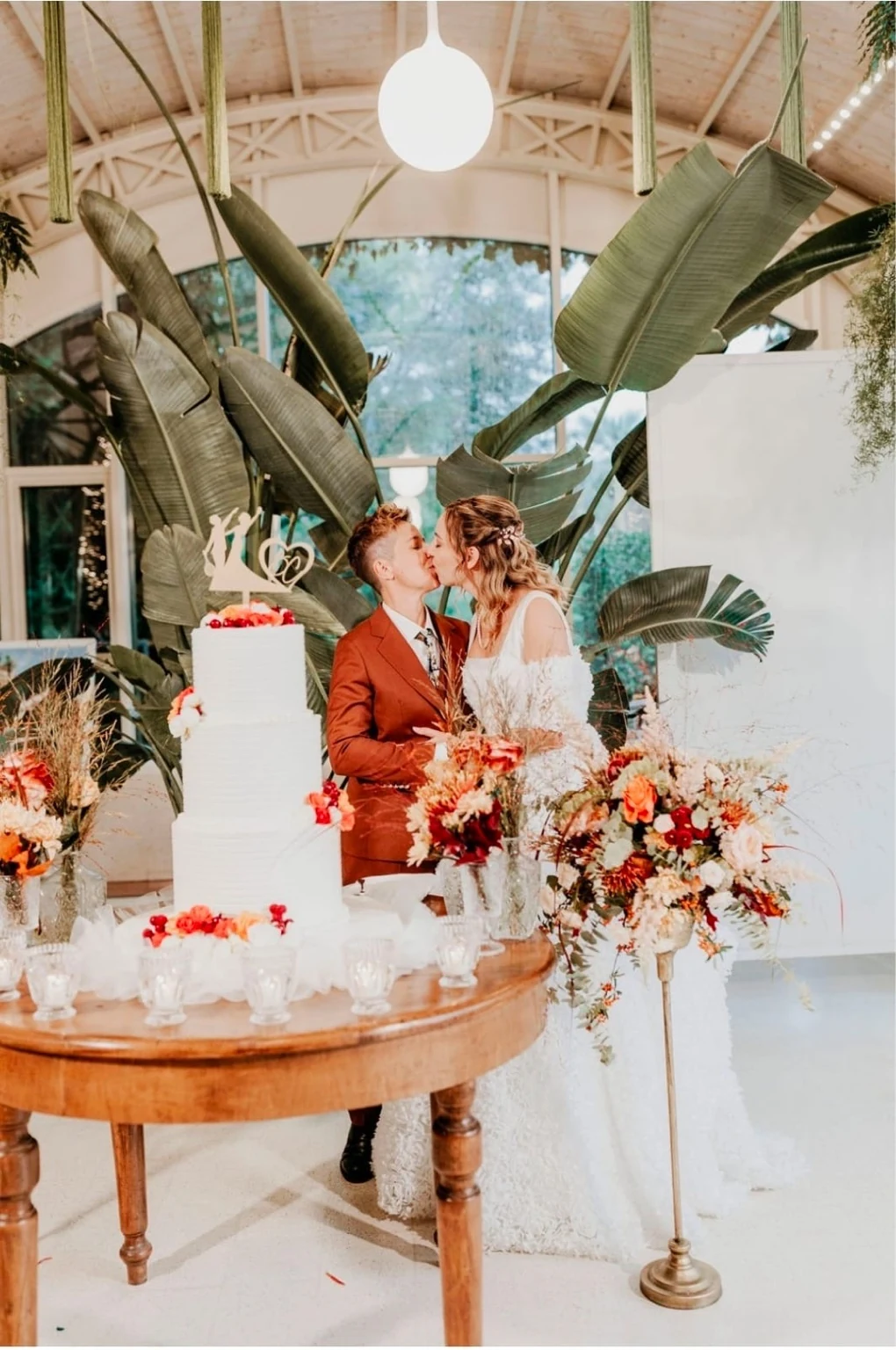 Claudia and Bruna kissing beside their wedding cake and large green leaves