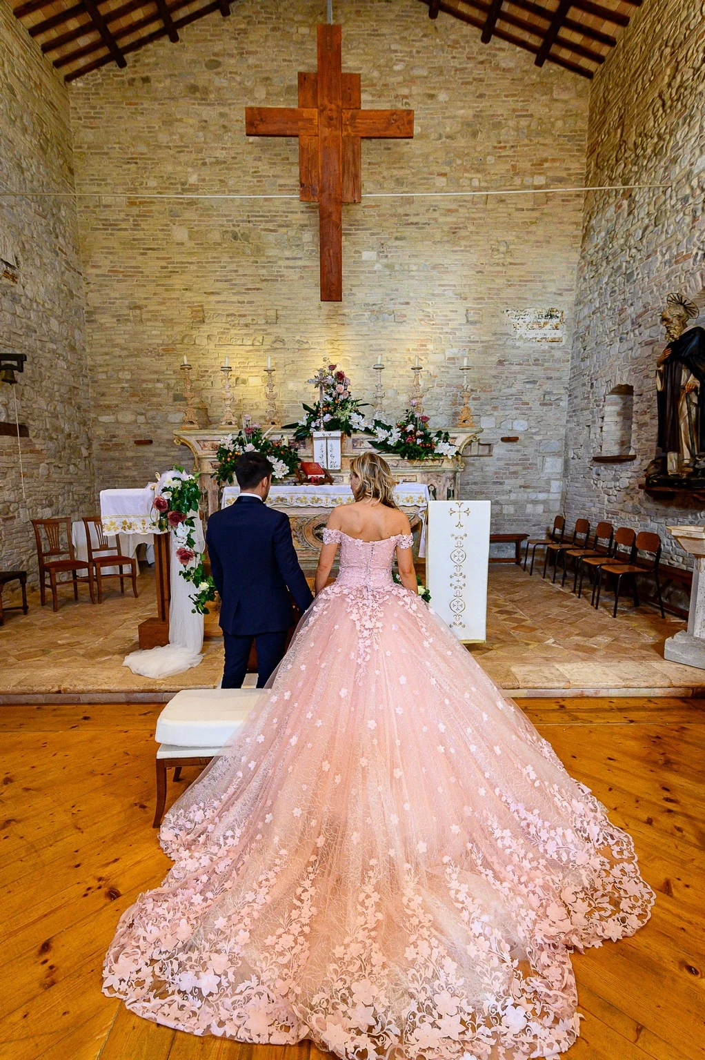 A couple standing at the altar of a stone church, with the bride’s pink dress and train in view