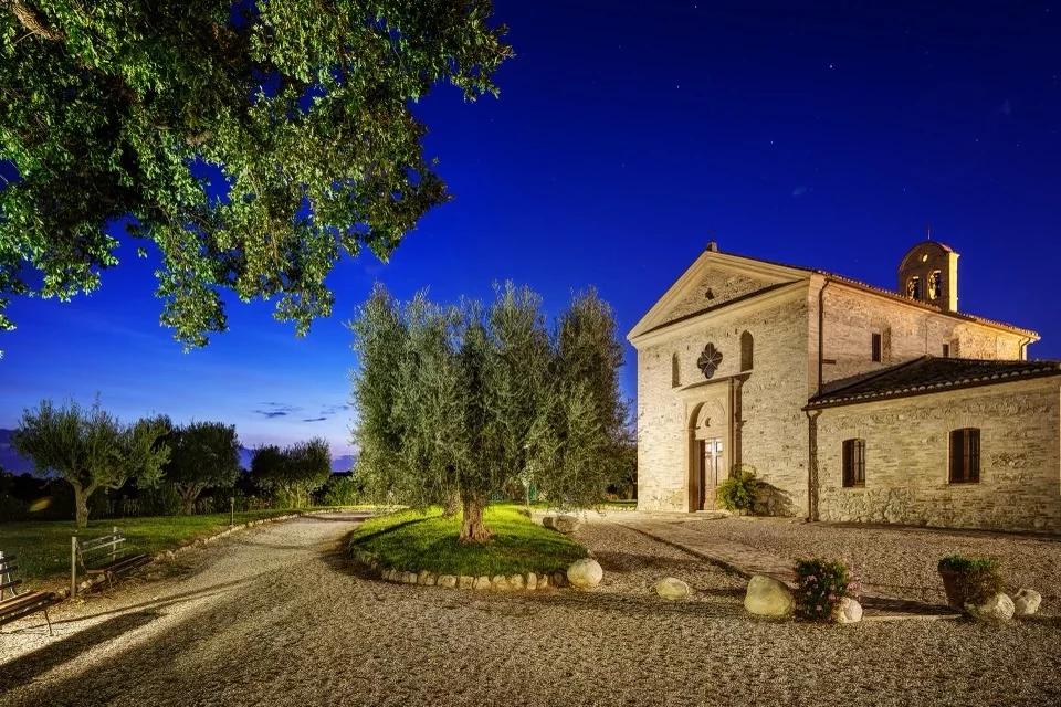 An illuminated stone church among olive trees beneath a blue evening sky