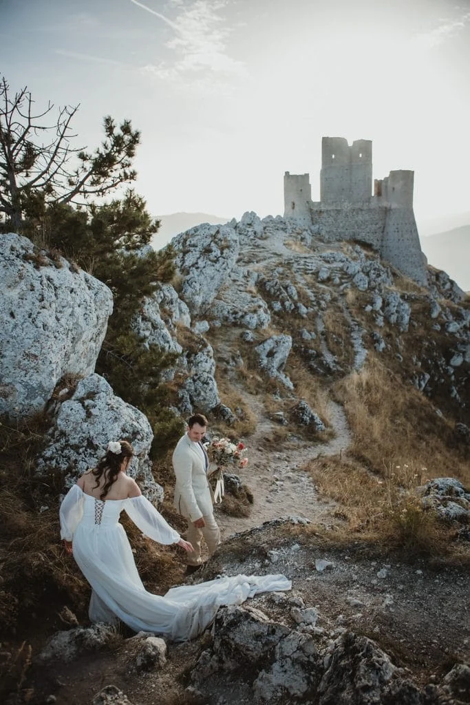 A wedding couple on a rocky path beneath a hilltop stone castle
